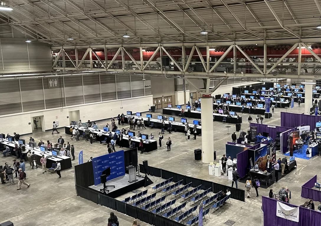 A convention hall floor with long rows of screens set up on draped tables, a theatre area and attendees walking through